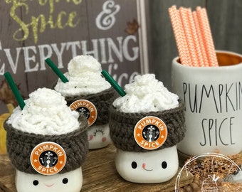 May include: A collection of handmade marshmallow-shaped mugs, each adorned with a crocheted brown cozy and a white whipped cream topping. The mugs feature a "Pumpkin Spice" sticker and a green straw. A mug with orange and white straws and a sign that says "Pumpkin Spice & Everything Nice" are also in the image.