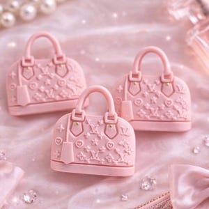 May include: Three light pink, dome-shaped handbags with handles and embossed floral patterns are arranged on a soft, pink surface. The scene includes a pearl necklace, diamonds, and a perfume bottle, creating a luxurious aesthetic.