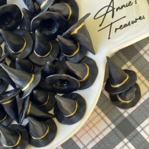May include: A collection of miniature black witch hats with gold trim, displayed on a white, scalloped-edge plate. The plate has the text "Annie's Treasures" in cursive. The background is a gray and white plaid pattern.