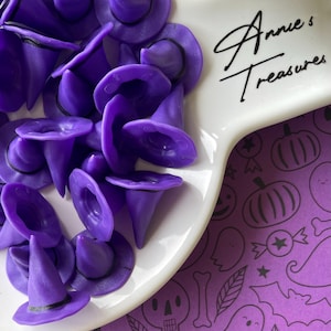 May include: A collection of small, purple witch hats with black bands, displayed on a white, curved dish. The dish is placed on a purple surface with Halloween-themed illustrations. The text "Annie's Treasures" is visible on the dish.