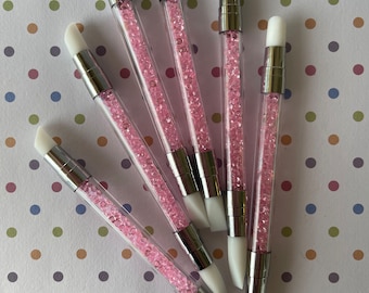 May include: Six white silicone nail art brushes with pink crystal handles. The brushes are arranged in a fan shape on a white background with pink, blue, and brown polka dots.