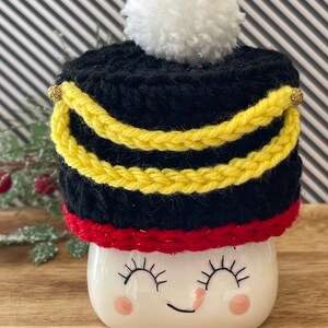 May include: A black and yellow crocheted hat with a white pom-pom on top, resting on a white mug with a smiling face. The hat has yellow trim and gold accents, and a red band at the base.