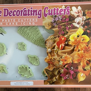May include: A box of cake decorating cutters with two layers of cutters. The box features a picture of flowers on the front and the text "Cake Decorating Cutters, Gum Paste Cutters for Cake Icing, This box contains two layers of cutters".