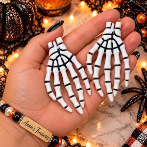 May include: A pair of white skeleton hand earrings with black detailing between the finger bones. The earrings feature a glossy finish and are shaped like human hand bones. The design includes a small attachment point at the top of each hand.
