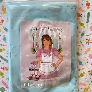 May include: A clear plastic bag filled with light blue material. A label on the bag features a woman in a pink apron and the text "Annie's Treasures". The label also includes the text "100 grams".