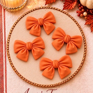 May include: Four solid orange bows with a knotted center detail arranged on a circular beige display board. The text "Annie's Treasures" is printed in a dark red script font at the bottom of the image, below the circular display board.