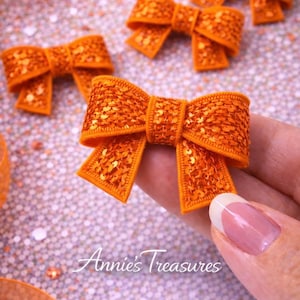 May include: Close-up of several orange sequined bows. The bows are arranged on a glittery surface, with one held by a hand. The bows have a textured appearance and are a vibrant orange color. The text "Annie's Treasures" is visible.