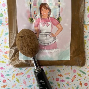 May include: A brown ice cream scoop with a black handle that reads "ICE CREAM" in white lettering, next to a scoop of brown ice cream. The scoop is in front of a bag with a label that reads "Annie's Treasures" with a cartoon image of a woman.