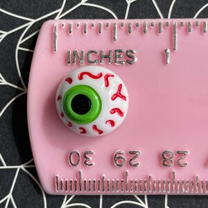 May include: A white googly eye with red veins and a green iris. The eye is approximately 1 cm in diameter and is sitting on a pink ruler with the words "INCHES" and "1" printed on it.
