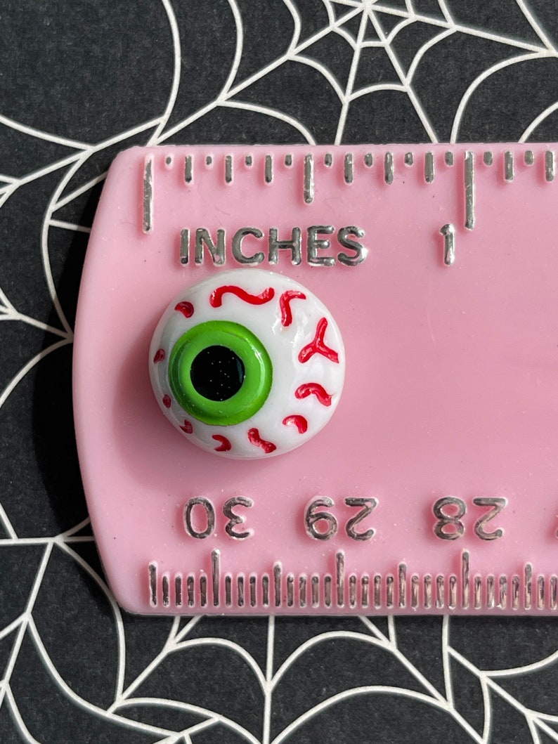 May include: A white googly eye with red veins and a green iris. The eye is approximately 1 cm in diameter and is sitting on a pink ruler with the words "INCHES" and "1" printed on it.