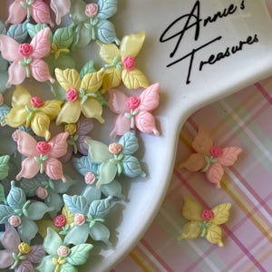 May include: Assortment of pastel colored butterfly shaped resin embellishments with a flower detail. The embellishments are displayed on a white plate with the text "Annie's Treasures" written on it.