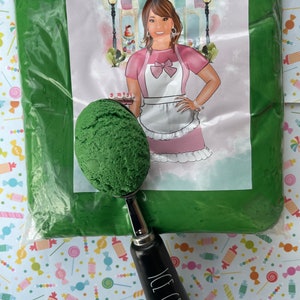 May include: A green plastic bag filled with green edible fondant. A green scoop of fondant is on a black ice cream scoop with the words "ICE CREAM" on the handle. The bag is sitting on a white background with a colorful pattern of candy.