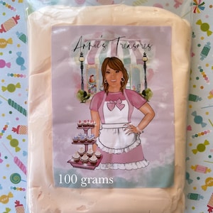 May include: A pink and white bag with a label that says "Annie's Treasures" and "100 grams". The label features a cartoon illustration of a woman wearing a pink apron and a bow, standing in front of a bakery display with cupcakes.