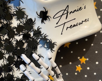 May include: A white, star-shaped dish filled with black spider-shaped confetti. A white skeleton hand is on the side of the dish. The dish has the text "Annie's Treasures" in black script. The background is black with white polka dots and yellow stars.