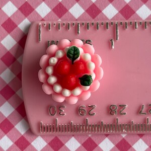 以下が含まれることがあります： 緑の葉で飾られた、白いフロスティングと赤いイチゴをトッピングしたミニチュアのピンクのケーキ。ケーキの直径は約2.5cmです。