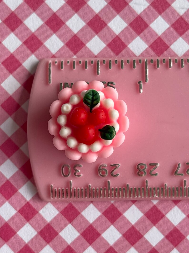 以下が含まれることがあります： 緑の葉で飾られた、白いフロスティングと赤いイチゴをトッピングしたミニチュアのピンクのケーキ。ケーキの直径は約2.5cmです。