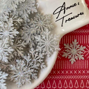 May include: A collection of silver glitter snowflakes on a white ceramic dish. The snowflakes are arranged on a red patterned surface. The dish has the text "Annie's Treasures" in black script.