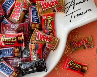 May include: A collection of miniature candy bar replicas, including Twix, Snickers, Kit Kat, and Reese's. The candy bars are arranged on a white ceramic dish with the words "Anne's Treasures" written on the side.