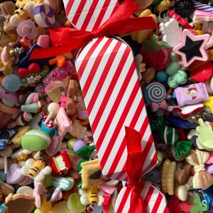 May include: A red and white striped candy-shaped box tied with a red ribbon, surrounded by a colorful assortment of miniature candies and sweets. The collection includes various shapes and colors, such as stars, strawberries, and bees.