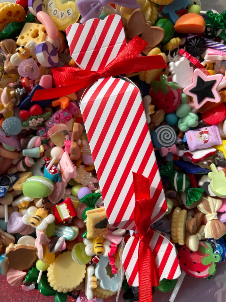 May include: A red and white striped candy-shaped box tied with a red ribbon, surrounded by a colorful assortment of miniature candies and sweets. The collection includes various shapes and colors, such as stars, strawberries, and bees.