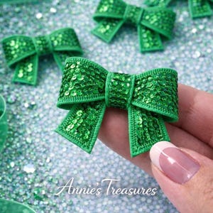May include: Close-up of several bright green sequined bows. The bows are arranged on a glittery surface, with one bow held in a hand. The bows have a classic bow shape with a center knot and two loops on each side. The text "Annie's Treasures" is visible.