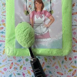 May include: A scoop of green ice cream in a black and silver ice cream scoop, with the words "ICE CREAM" printed on the handle. The scoop is in front of a bag of green slime and a printed label.