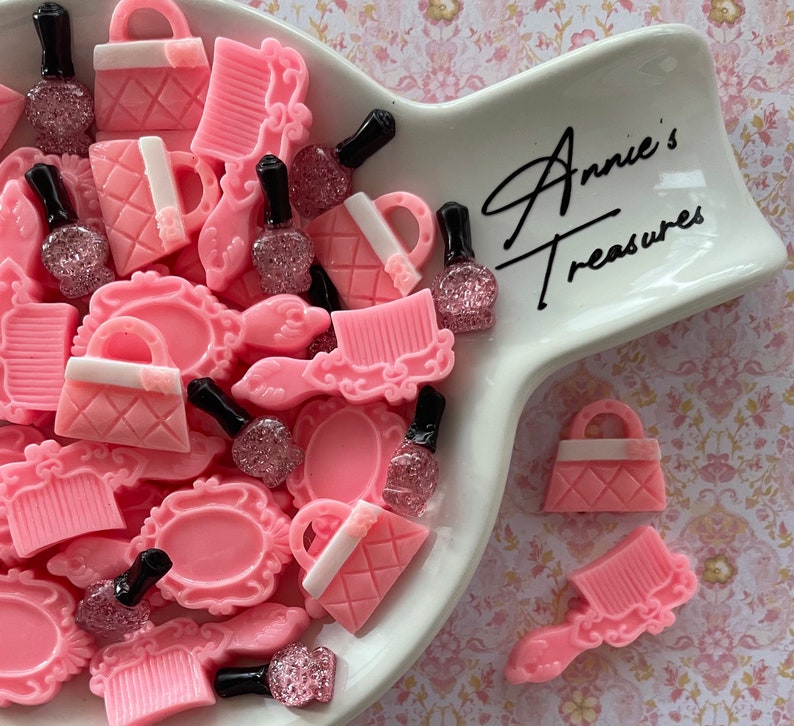 May include: A white ceramic dish with pink and white resin charms in the shape of purses, combs, and perfume bottles. The dish has the text "Annie's Treasures" written on it.
