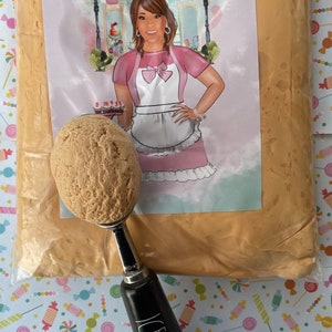 May include: A clear plastic bag filled with light brown product, with a scoop of the same product in a black and silver ice cream scoop. The bag has a label with the text "Annie's Treasures" and a cartoon illustration of a woman.