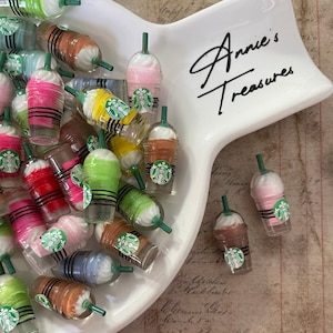 May include: A white ceramic dish with the text "Annie's Treasures" in black script. The dish is filled with miniature plastic cups with a variety of colors and designs, resembling Starbucks Frappuccinos.