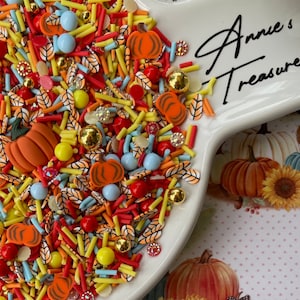 May include: A white ceramic dish filled with colorful sprinkles, including orange, yellow, red, blue, and brown. The sprinkles are shaped like pumpkins, leaves, and sticks. The dish has the text "Annie's Treasure" written on it.