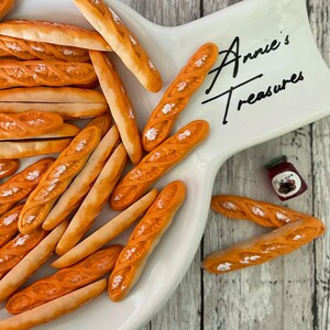 May include: A white ceramic serving dish with a brown wooden background. The dish is filled with miniature, orange-colored, baguette-shaped bread. The dish has the text "Annie's Treasures" written on it.