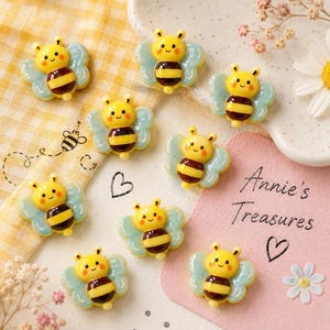 May include: A collection of yellow, black, and brown bee-shaped embellishments with light blue wings. Each bee has a smiling face and is decorated with black stripes. The background includes a pink card with the text "Annie's Treasures" and a heart symbol.