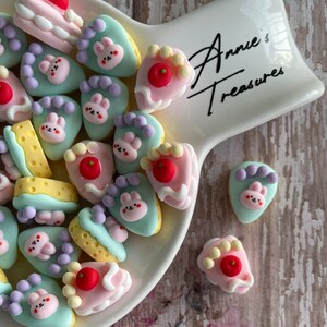 May include: A white ceramic dish filled with colorful miniature cake charms. The charms are shaped like slices of cake with different toppings, including cherries, sprinkles, and bunny faces. The dish has the text "Annie's Treasures" written on it.