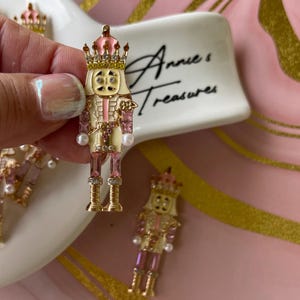 May include: A decorative nutcracker charm with a pink and gold crown, pink and gold accents, and faux pearls. The charm is held in front of a white dish with the text "Annie's Treasures" in black script. Other nutcracker charms are in the background.