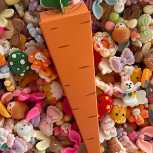 May include: An orange paper carrot with green leaves is surrounded by a variety of colorful resin charms. The charms include bows, bunnies, donuts, and other cute shapes. The overall theme is Easter and spring.