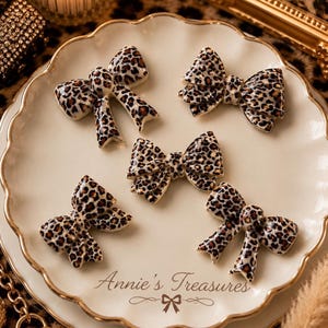 May include: Five decorative bows with a leopard print pattern are arranged on a cream-colored plate with a gold rim. The bows are brown and black. The plate has the text "Annie's Treasures".