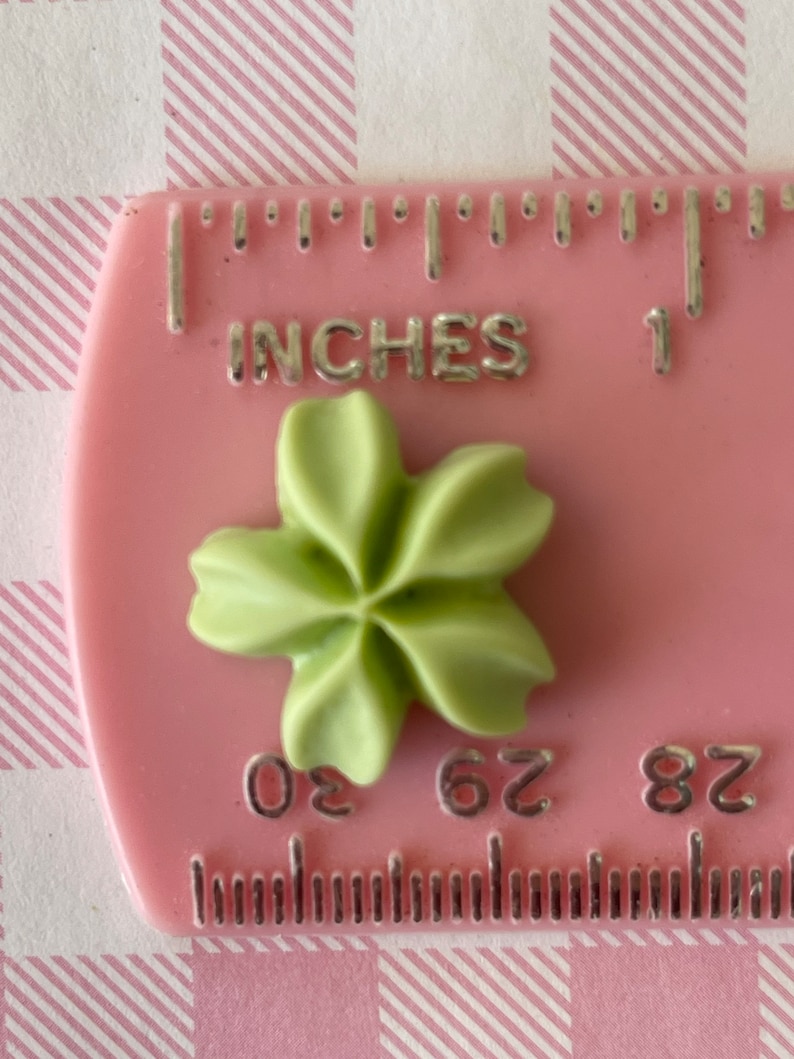 May include: A green five-pointed star-shaped embellishment, approximately 0.625 inches in diameter, is shown on a pink ruler with the word "INCHES" and the numbers "0", "62", and "82" visible.