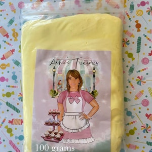 May include: A clear plastic bag containing a block of yellow material. A label on the bag features a cartoon illustration of a woman in a pink dress and apron, with the text "Annie's Treasures" and "100 grams". The background is a colorful candy-themed pattern.