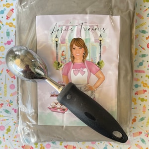 May include: A shiny, stainless steel ice cream scoop with a black handle rests on a package. The package features a colorful illustration with the text "Annie's Treasures". The background has a candy-themed pattern.
