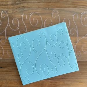 May include: A light blue silicone baking mat with a raised swirl pattern. The mat is on top of a clear plastic sheet with the same pattern.