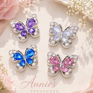May include: Four jeweled butterfly brooches in purple, blue, pink, and clear. Each butterfly has a silver-colored frame and is adorned with sparkling crystals. The image includes the text "Annie's Treasures".