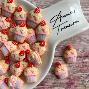 May include: A close-up of a white ceramic serving dish with a pink and yellow cupcake charm. The dish has the text "Annie's Treasures" written on it. The cupcake charm has a pink frosting, a red strawberry, and colorful sprinkles.