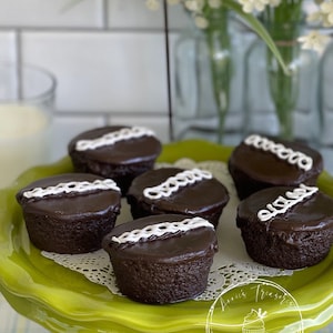 May include: Six chocolate cupcakes with white frosting and a decorative design on a green cake stand. The cupcakes are on a white doily. The stand is on a plaid tablecloth. The text "Annie's Treasures" is visible on the stand.