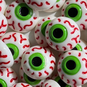 May include: A close-up of several white eyeball-shaped decorations with green pupils and red veins. The decorations are made of a smooth, glossy material and have a realistic appearance.