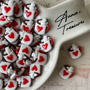 May include: A white ceramic bowl filled with small, white ceramic mugs with red hearts and brown tops. The mugs are arranged in a random pattern. The bowl is on a white surface with the text "Annie's Treasures" written in black.
