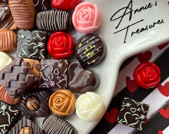 May include: A white plate with a variety of chocolate candies and decorative roses. The plate is on a black and white striped background with red hearts. The plate has the text "Annie's Treasures" written on it.