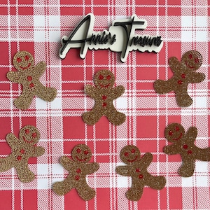May include: A collection of gingerbread man ornaments with red accents and a glittery gold finish, arranged on a red and white plaid background. The image includes the text "Annie's Treasure" in a black and white design.