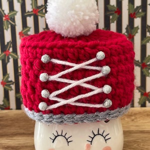 May include: A white ceramic mug with a red crocheted hat with a white pom pom on top. The hat has white and silver accents. The mug has a smiling face with eyelashes.