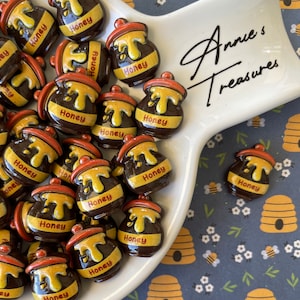May include: A white ceramic dish filled with small, brown, honey pot-shaped charms. Each charm has a yellow honey drip and the word "Honey" written on it. The dish is on a blue background with a beehive pattern.