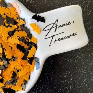 May include: A white ceramic dish filled with black and gold bat-shaped confetti. The dish has the text "Annie's Treasures" written on it.